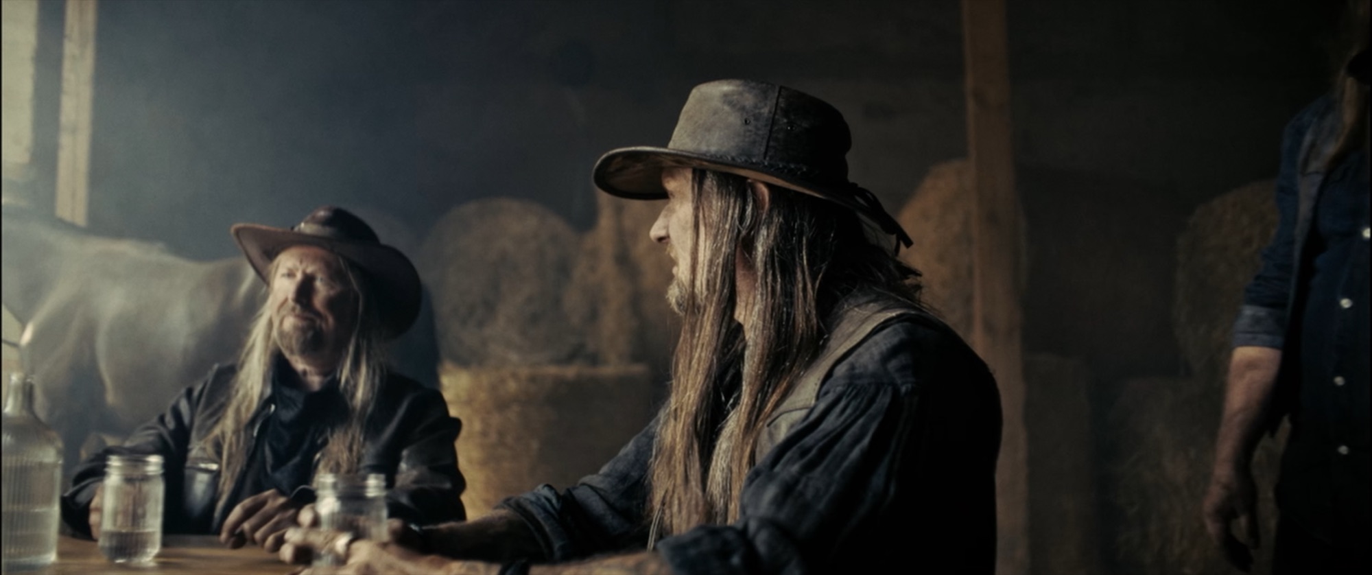 Two bearded, long-haired men in hats sitting at a table with mason jars in a smoky barn - Amon Amarth